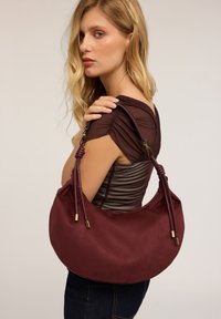 Sac à épaule bordeaux avec une texture lisse, en forme de croissant, et des poignées nouées ajustables avec des accents en ton or.