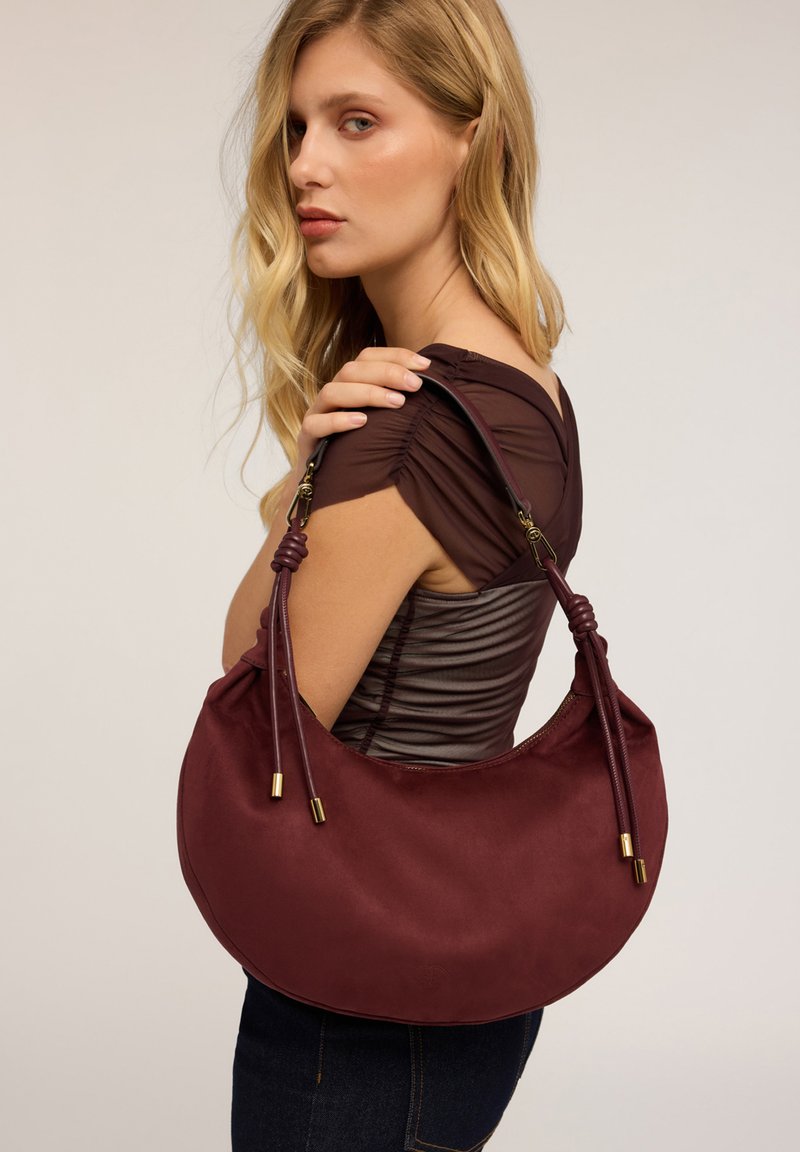 Sac à épaule bordeaux avec une texture lisse, en forme de croissant, et des poignées nouées ajustables avec des accents en ton or.