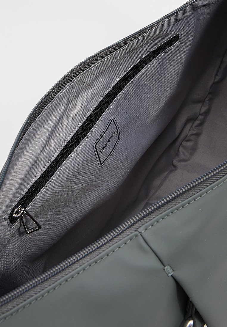 Interior de bolsa gris que muestra un bolsillo con cremallera de color negro y una etiqueta de tela cosida en el forro.