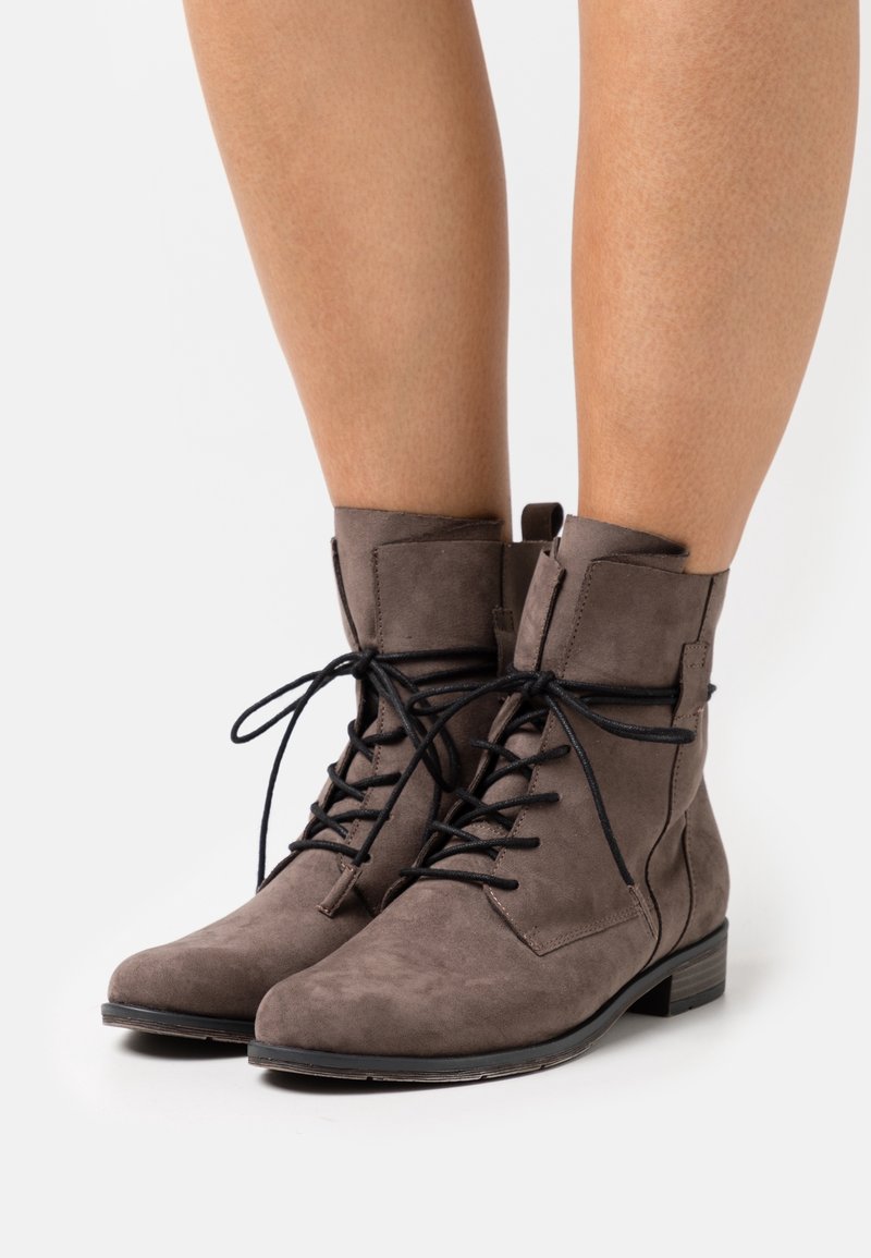 Bottes en daim taupe montant à la cheville avec des lacets noirs, portées sur des jambes nues sur un fond blanc uni.