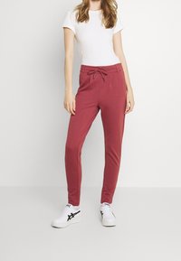 Pantalons de jogging ajustés rouges avec une taille à cordon, présentant une texture lisse et un design fuselé. Associés à un t-shirt blanc uni et des chaussures blanches.