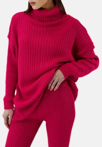 Fuchsia ribbad stickad tröja med hög polokrage och oversized passform, i kombination med matchande ribbade leggings. Mjuk textur och avslappnad design.