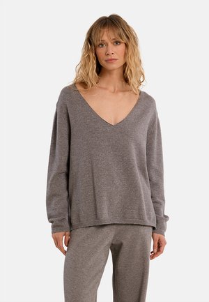 Donna con capelli biondi mossi che indossa un maglione grigio ampio con scollo a V e pantaloni grigi abbinati su uno sfondo uniforme.
