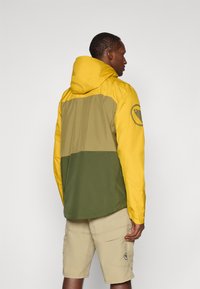 Farbblockierte Jacke mit einer gelben Kapuze und oberem Abschnitt, olivgrüner Mitte und dunklem grünen unteren Abschnitt; mit Logo auf dem Ärmel.