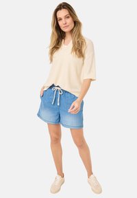 Beżowy sweter z dzianiny z dekoltem w serek, jasnoniebieskie szorty dżinsowe na sznurku oraz kremowe sneakersy. Luźny krój i miękka tekstura w swobodnym stroju.