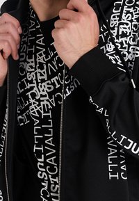 Czarna bluza z kapturem na zamek błyskawiczny z białym wzorem tekstu. Miękka tkanina, ściągacze przy mankietach i kaptur ze sznurkiem. Nosi się ją na czarnej koszulce z podobnym wzorem tekstu.