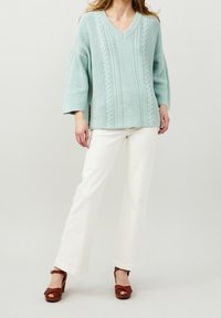 Femme portant un pull en maille torsadée oversize bleu clair, un pantalon blanc à jambes larges et des sandales à talons hauts marron, debout devant un fond uni.