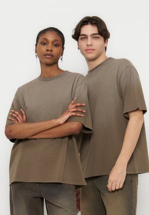 Δύο νέοι ενήλικες φορούν φαρδιά μπεζ μπλουζάκια και καφέ παντελόνια, στέκονται μπροστά σε λευκό απλό φόντο.