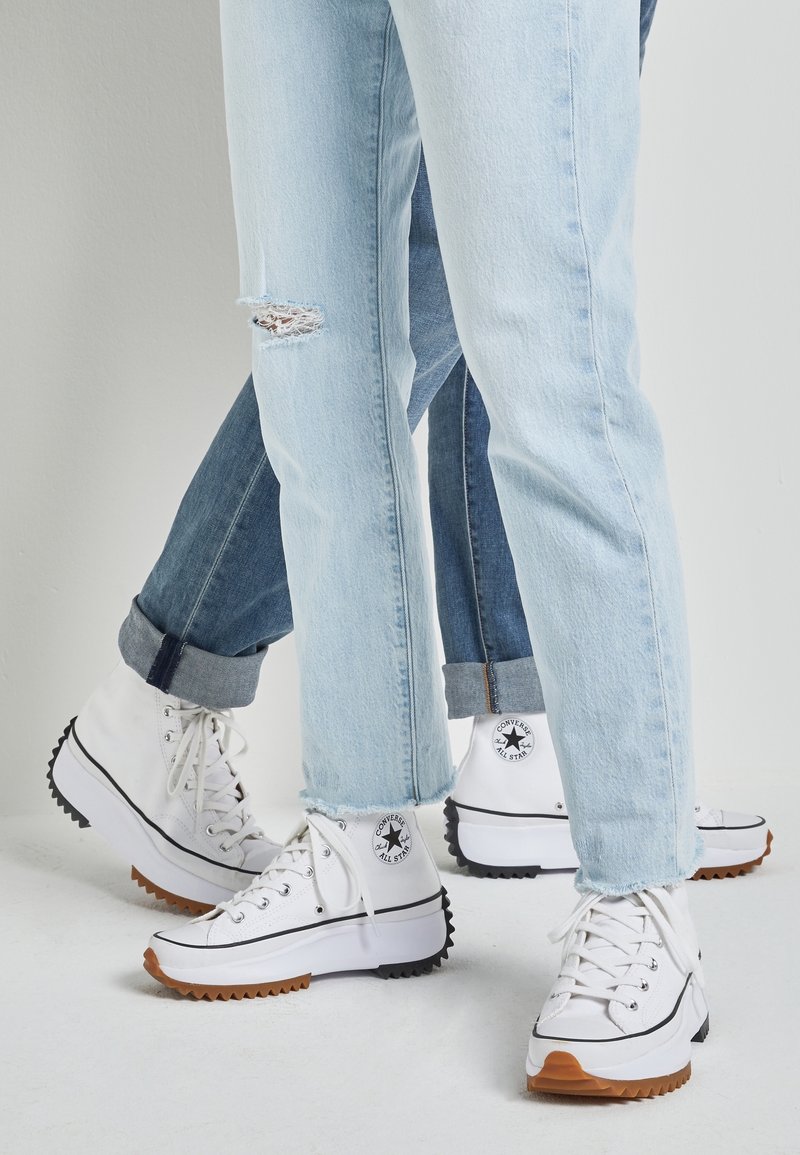Baskets montantes blanches avec des accents noirs, présentant une semelle en caoutchouc texturée. Portées avec un jean en denim bleu clair et un jean en denim bleu foncé, l'une des paires a une déchirure.