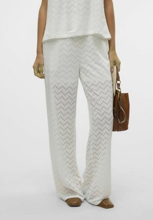 Witte wijde broek met een zigzagpatroon, met perforaties voor textuur. Gecombineerd met een casual top en een bruine tas.