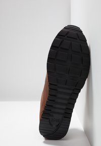 Chaussure de sport marron avec une tige texturée et une semelle extérieure en caoutchouc noir présentant des motifs de bande de roulement proéminents. Le logo de la marque est visible sur la semelle.