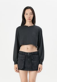 Svart kort sweatshirtt med lange ermer og en subtil logo på brystet, sammen med et svart denimskjørt som har forlommer.