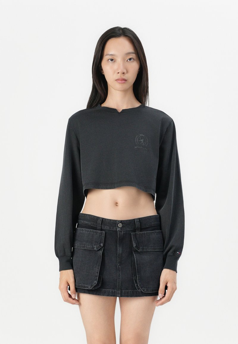 Svart kort sweatshirtt med lange ermer og en subtil logo på brystet, sammen med et svart denimskjørt som har forlommer.