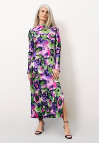 Lange bloemenjurk in groen, paars en pink. Heeft lange mouwen, een hoge nek en een zijsplits, gemaakt van een gladde stof. Zwarte hakken.