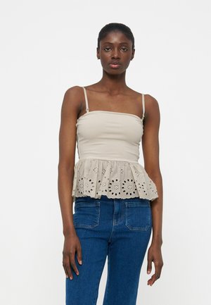 Ung kvinne iført en beige ermeløs topp med hullmønster peplum og blå jeans, stående foran en ensfarget hvit bakgrunn.