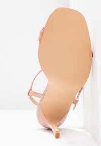 Chaussure à talon haut nude avec brides, bout pointu et talon fin. Finition en cuir verni lisse et semelle en caoutchouc texturée.