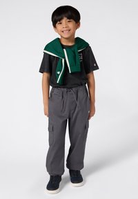 Młodzieżowy strój składający się z czarnej koszulki, szarych spodni cargo z bocznymi kieszeniami oraz zielonego swetra zarzuconego na ramionach. Czarne sneakersy.