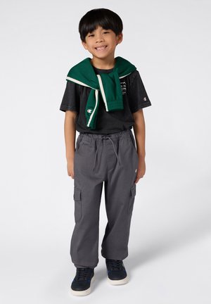 Młodzieżowy strój składający się z czarnej koszulki, szarych spodni cargo z bocznymi kieszeniami oraz zielonego swetra zarzuconego na ramionach. Czarne sneakersy.