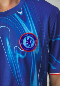 Plava nogometna majica sa vrtložnim grafičkim dizajnom, koja ima kružni logo Chelsea nogometnog kluba u centru, vidljivi detalji teksture.