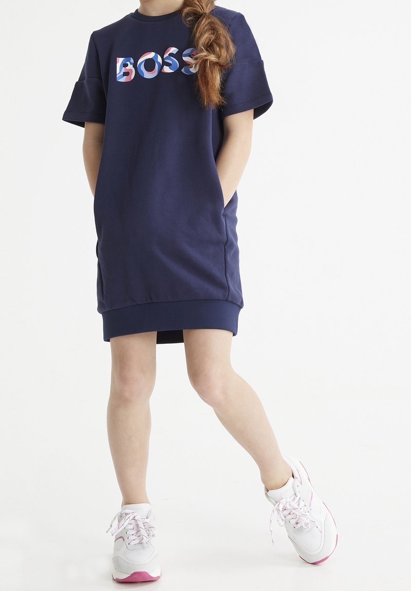 Enfant debout, les mains dans les poches, portant une robe bleu marine avec le logo "BOSS" et des baskets blanches avec des accents roses.