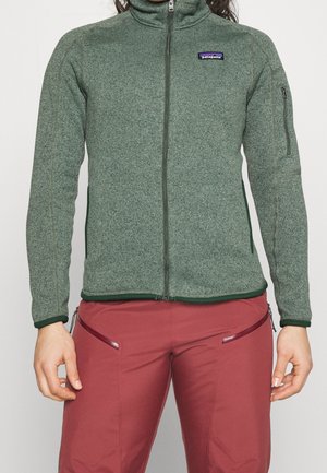 Femme portant une polaire zippée verte Patagonia avec une poche zippée sur la manche et un pantalon rouge avec des poches zippées.