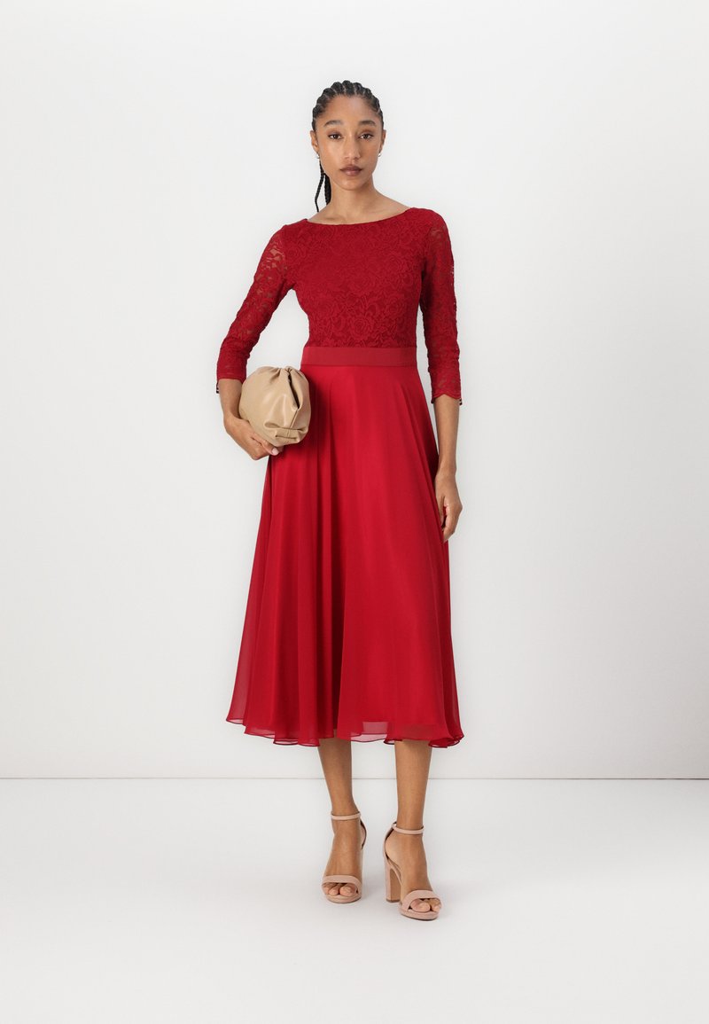Rotes Kleid mit Spitzenoberteil und Dreiviertelärmeln, fließendem Rock und festem Taillenbereich. Eine beige Clutch und helle Absätze runden den Look ab.