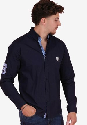 Homme portant une chemise bleu marine à boutons avec un col intérieur à carreaux bleus et un écusson sur la poitrine gauche et la manche, les mains dans les poches, regardant vers la droite.