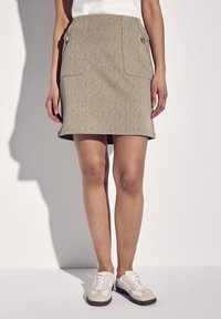 Jupe crayon kaki avec poches latérales, fabriquée en tissu texturé. Associée à des baskets blanches avec des accents beige et des semelles marron.