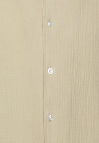 Camicia in tessuto beige con texture, dotata di una fila di bottoni verticale e piccoli bottoni bianchi rotondi.