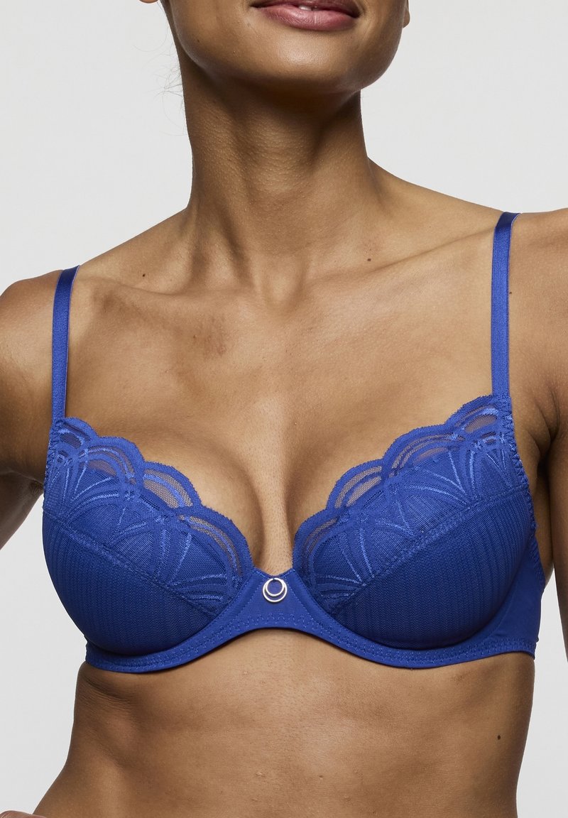 Koningsblauwe bh met kanten rand, beugelondersteuning en gegolfde randen. Heeft een decoratief metalen accent in het midden. Gladde textuur.