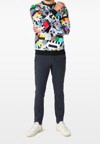 Bunt gemusterter Sweatshirt mit geometrischen Designs in Rot, Blau, Grün und Gelb; kombiniert mit dunkel strukturierten Hosen und weißen Sneakers.