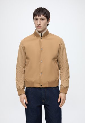 Mann trägt eine beige Bomberjacke mit Knöpfen, gerippten Bündchen und Kragen, kombiniert mit dunkelblauen Jeans, steht vor einem schlichten weißen Hintergrund.