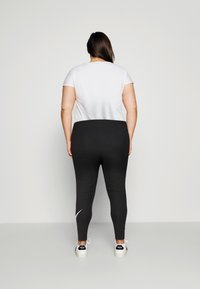 Kvinna med långt mörkt hår står vänd mot en vanlig ljus vägg, iklädd en vit kortärmad skjorta, svarta leggings och vita sneakers.