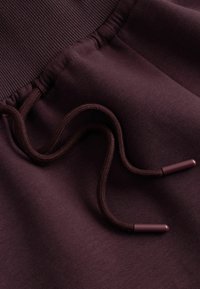 Gros plan d’un pantalon de survêtement bordeaux avec une ceinture côtelée et un cordon avec embouts en plastique.