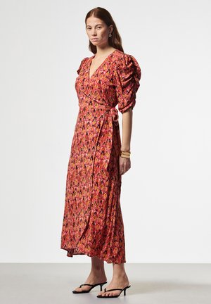 Vrouw die een roze en oranje gewatteerde wikkeljurk met pofmouwen, sandalen en een gouden armband draagt, staand tegen een effen achtergrond.