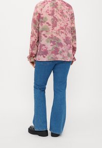 Femme portant un chemisier rose à manches longues à motifs floraux, un jean évasé bleu et des chaussures à plateforme noires, debout sur un fond blanc uni.