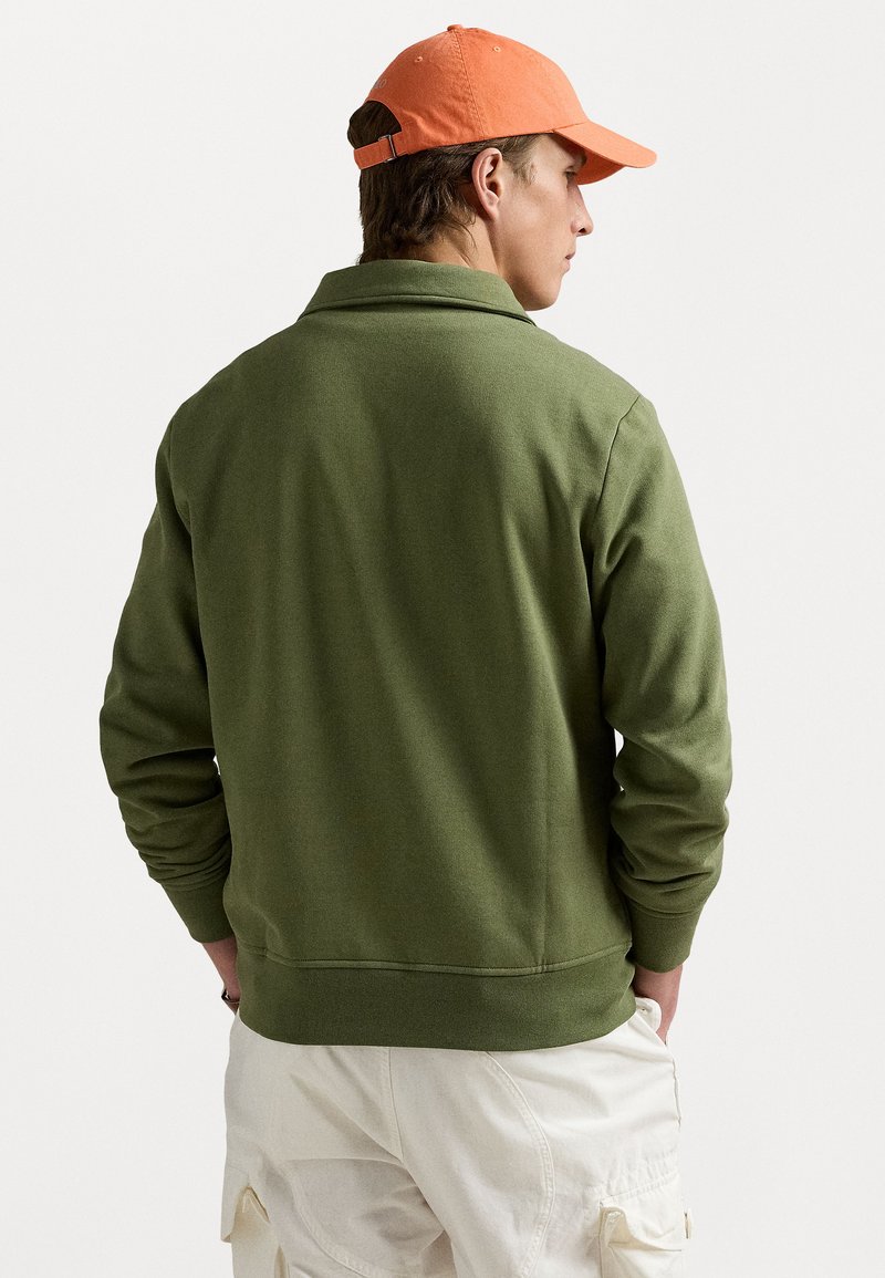 Olijfgroene sweatshirt met een kraag en geribbelde manchetten, gedragen met lichtgekleurde cargobroeken en een oranje pet.