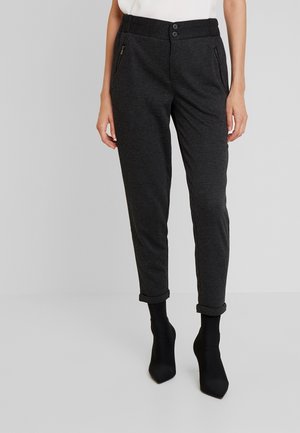 Femme portant un pantalon slim gris foncé avec revers roulés, poches zippées, et bottines noires à bout pointu, debout sur un sol gris clair.