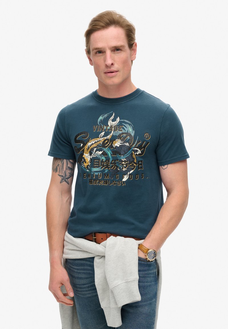 Camiseta de algodón de manga corta en azul oscuro con un estampado gráfico en elementos dorados, azules y negros. Llevada con jeans azules y una sudadera gris atada a la cintura.