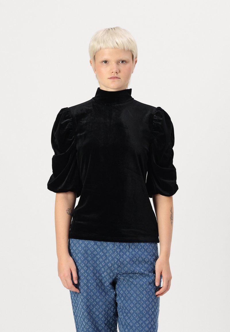 Mustmustagu musta sametist top, millel on kõrge kaelus ja puhvis varrukad. Omab siledat tekstuuri ja elegantset siluetti, koos mustriliste siniste pükstega.