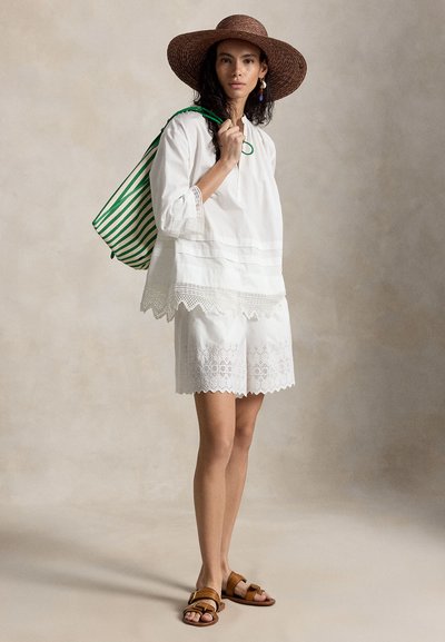 Γυναίκα που φοράει λευκό μπλουζάκι με δαντελένιο τελείωμα και σορτς, καφέ καπέλο με φαρδύ γείσο, καφέ σανδάλια και κρατάει πράσινη και λευκή ριγέ τσάντα tote.