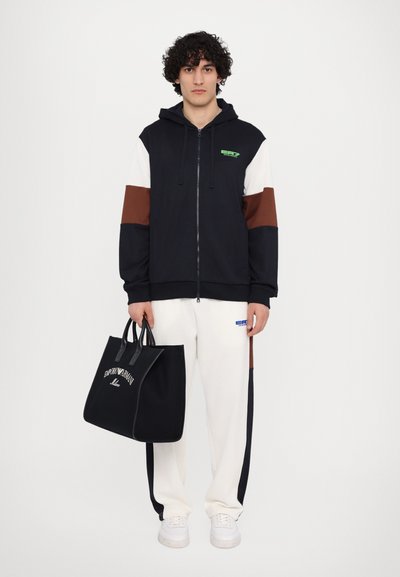Felpa con cappuccio e zip in blu navy, bianco e marrone, abbinata a pantaloni crema. Il modello tiene una borsa tote nera con logo bianco. Sneakers bianche.