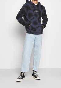 Felpa con cappuccio tie-dye blu navy con tasca frontale e cappuccio regolabile, abbinata a jeans a gamba larga blu chiaro e sneakers nere alte.