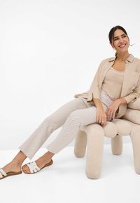 Lichtbeige linnen overhemd over een beige tanktop, gecombineerd met gestructureerde lichtgrijze broeken. Witte slidesandalen met een kruisbandontwerp.