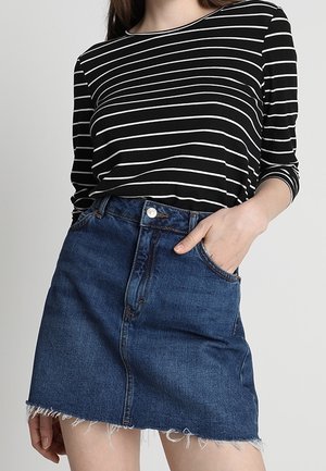 Femme portant une chemise à manches longues rayée noir et blanc, rentrée dans une jupe en jean bleue à ourlet effiloché, avec une main dans la poche.