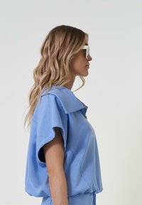 Gaiši zils, īsām piedurknēm, brīvi pieguļošs tops ar apkakli un rāvējslēdzēja aizdari, izgatavots no mīksta auduma. Gluda tekstūra ar smalkām šuvēm.