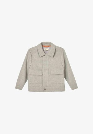 Ung dreng med bølget brunt hår iført en lysegrå jakke over en hvid skjorte, stående mod en ensfarvet beige baggrund.