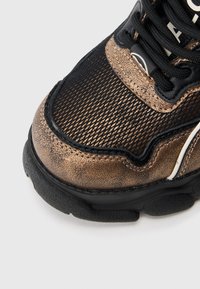 Μαύρο και μεταλλικό μπρούτζινο αθλητ ικό παπούτσι με υφή στην επιφάνεια, τμήματα από δίχτυ και μαύρα κορδόνια. Παχύ ελαστικό πάτο με διακριτικό λογότυπο.