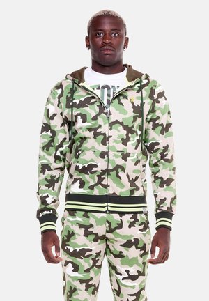 Uomo adulto in piedi, indossa una felpa con cappuccio mimetica verde, marrone e beige con zip e pantaloni coordinati, con una t-shirt bianca con grafica sotto.