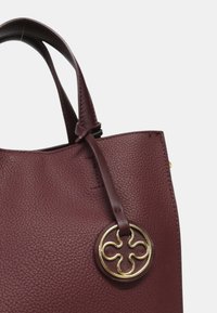 Sac à main en cuir texturé marron foncé avec doubles poignées et un charme décoratif rond en or attaché près du bord supérieur.